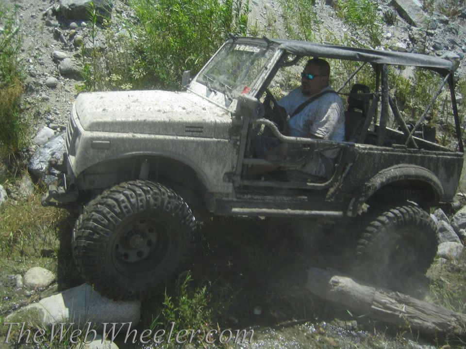 Muddy off-road vehicle driving through water and over a log. Driver wears sunglasses