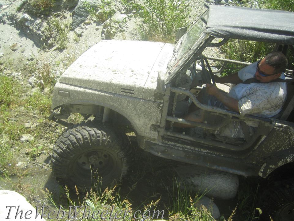 Mud-covered 1987 Suzuki Samurai with a driver, off-roading in a rocky, overgrown area