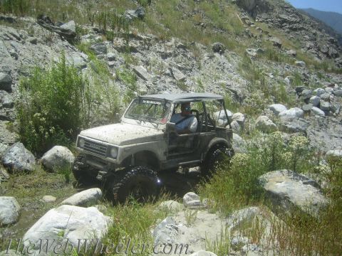 1987 Suzuki Samurai - A white 1987 Suzuki Samurai navigates a challenging off-road course with rocky terrain and dense vegetation.