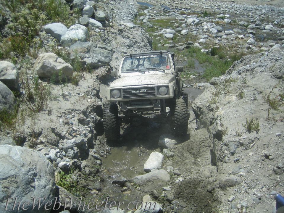 White 1987 Suzuki Samurai off-roading through a rocky, muddy stream bed
