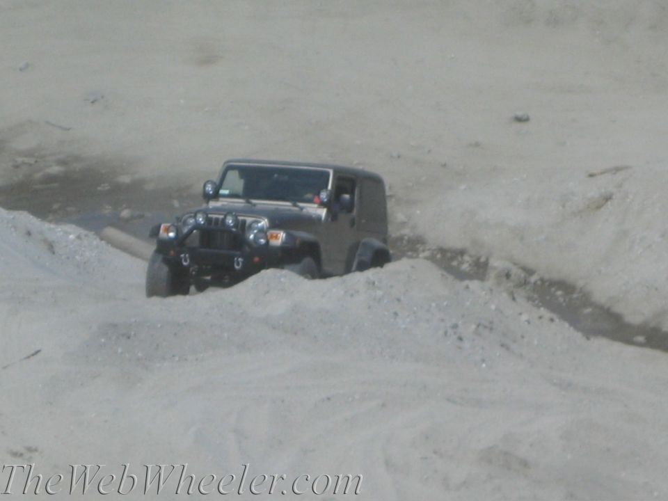 Dark-colored Jeep Wrangler navigating a sandy, uneven terrain with auxiliary lights on the front bumper