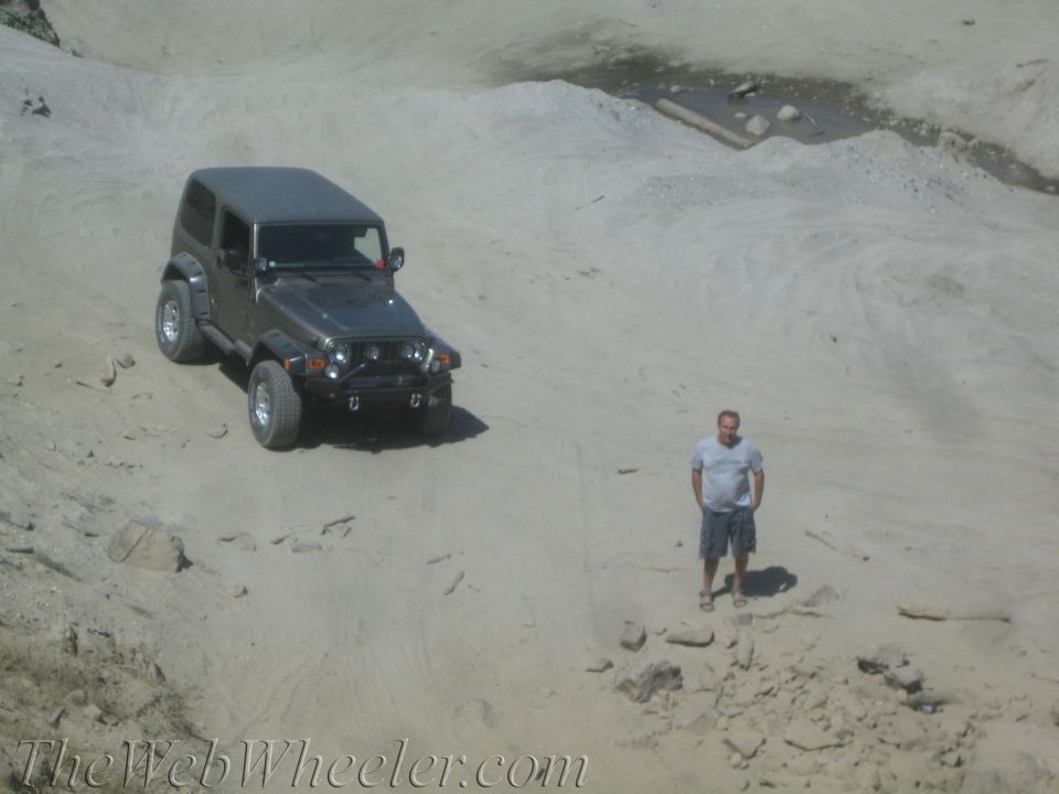 Man standing next to a dark gray Jeep Wrangler on a sandy, off-road terrain