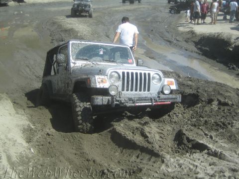 1987 Suzuki Samurai - A silver Jeep Wrangler TJ struggles through a muddy off-road course, with other vehicles and spectators visible in the background.