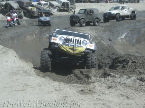 1987 Suzuki Samurai - A heavily modified Jeep Wrangler navigates a challenging muddy terrain, with other vehicles and spectators present.