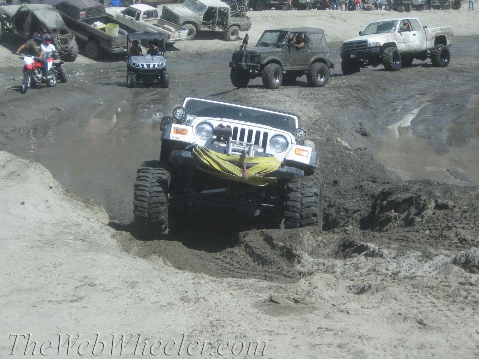 White Jeep Wrangler with big tires going up a muddy hill. Other off-road vehicles and people in background