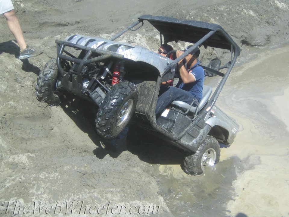 Silver side-by-side utility vehicle with black roof and roll cage, tilted on a muddy incline with two occupants