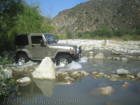 1987 Suzuki Samurai - Jeep Wrangler TJ navigates a shallow river with rocky terrain and hills in the background.