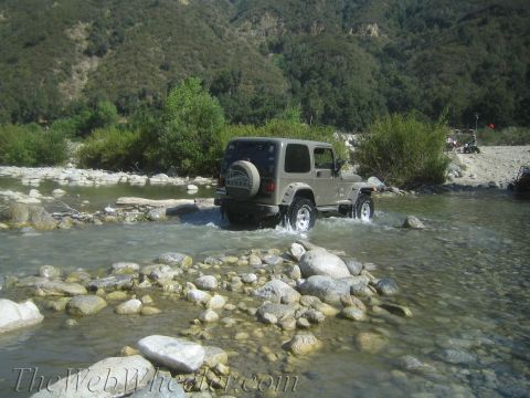 1987 Suzuki Samurai - A tan Jeep Wrangler navigates a shallow, rocky riverbed with trees and mountains in the background.