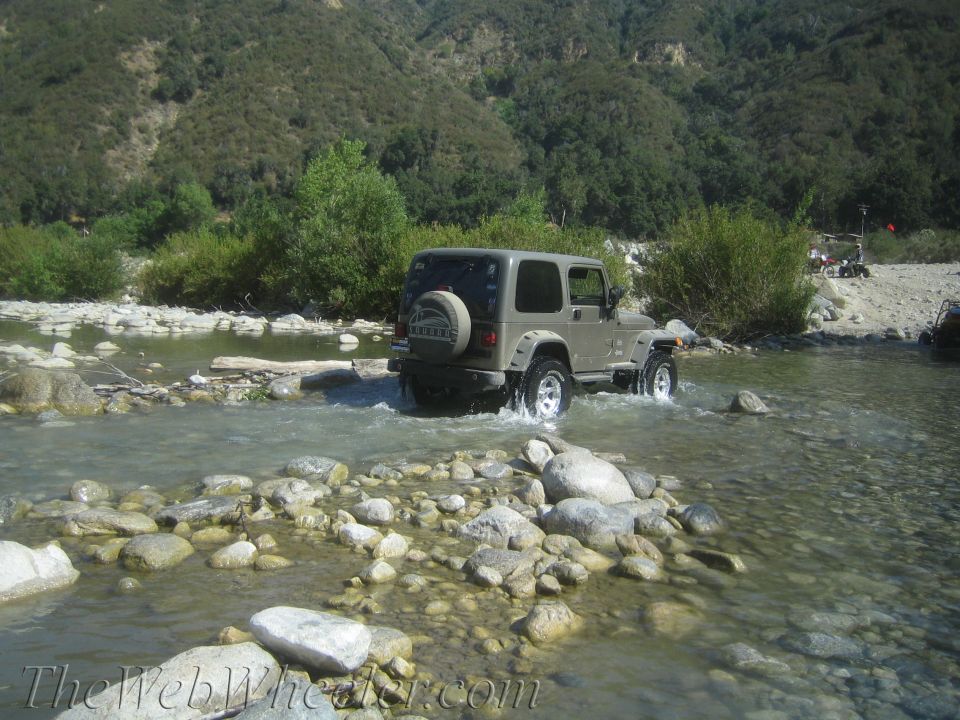 Tan Jeep Wrangler driving through a rocky river, spare tire on back