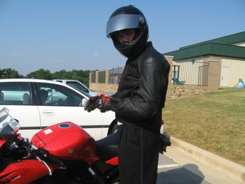 1988 Ford Bronco - A person in motorcycle gear stands beside a red motorcycle with a white car in the background.