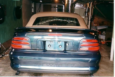 1991 Ford LTD Crown Victoria - Green Ford Mustang GT convertible with tan soft top, photographed from the rear in a basement garage; Mustang GT badge visible.