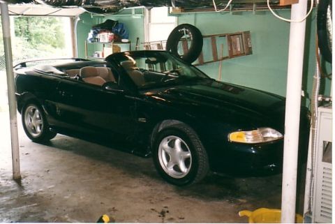1991 Ford LTD Crown Victoria - Passenger-side front three-quarter view of a black SN95 Ford Mustang GT convertible with the top down, parked in a green-walled carport wit…
