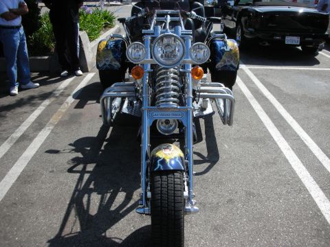 1998 Ford Crown Victoria - A custom trike motorcycle with a prominent front wheel and chrome accents is parked in a lot next to a black convertible.