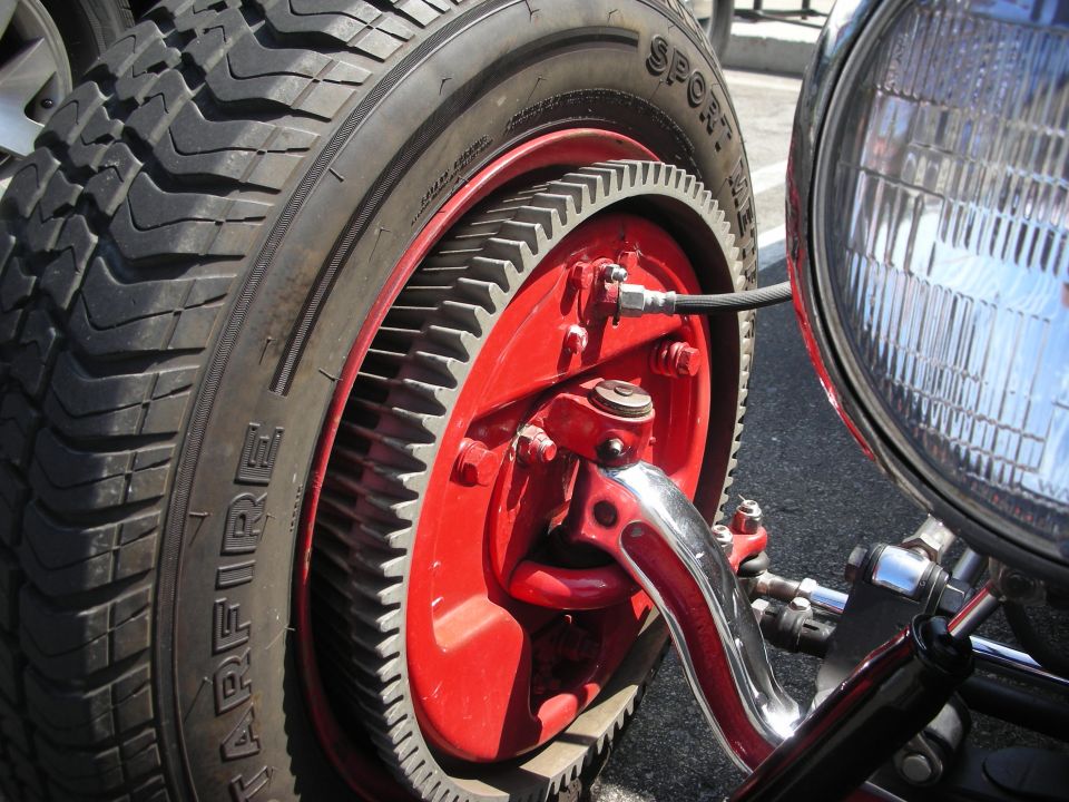 Close-up of a red wheel with a large gear ring, a Firestone Sport King tire, and a chrome headlight on a custom car