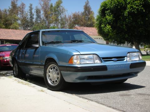 1998 Ford Crown Victoria - Blue 1987 Ford Mustang parked curbside with a red car visible behind it.