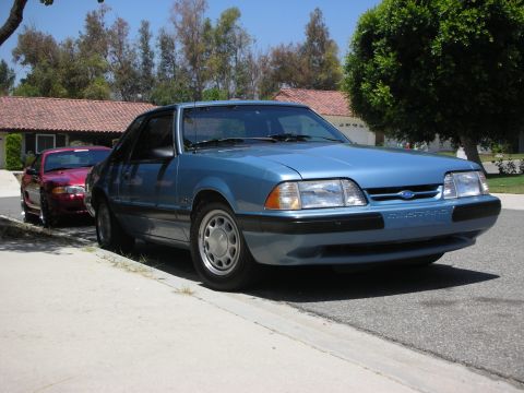 1998 Ford Crown Victoria - A blue Ford Mustang is parked on the street next to a sidewalk, with another car behind it.