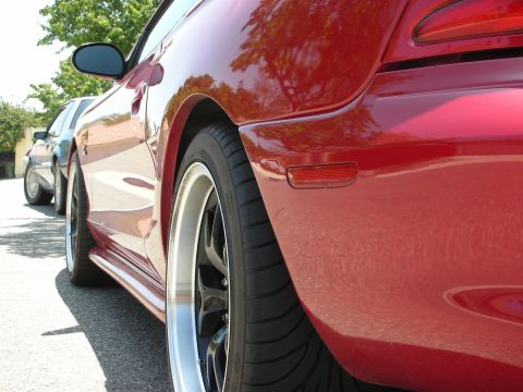 1998 Ford Crown Victoria - Close-up of a red sports car's rear quarter panel, custom wheels and a side skirt, with another vehicle parked nearby.