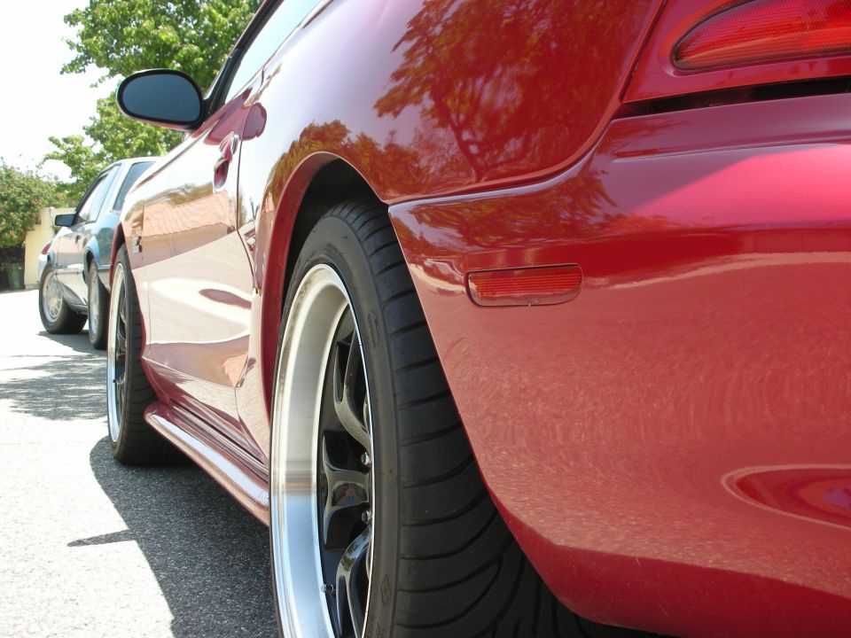 Red sports car rear quarter panel with aftermarket wheels and side skirt. Another car is visible in the background