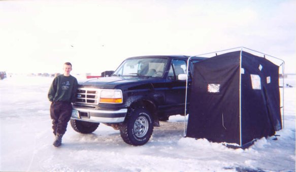 Black lifted Ford SUV on snowy ice with man leaning on fender and dark ice-fishing tent