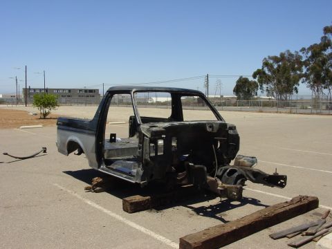 1994 Ford Bronco - A stripped full-size Ford truck body shell resting on wood blocks in an empty asphalt parking lot, doors, glass, engine, front clip and int…