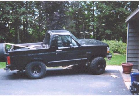 1996 Ford Bronco - A black Ford Bronco with its removable hardtop roof off, shown in side profile on a paved driveway next to a gray house. It sits on black s…