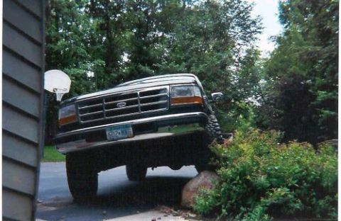 1996 Ford Bronco - A lifted dark Ford Bronco flexes its suspension with the passenger front wheel climbed up on a large boulder at the edge of a driveway, fro…