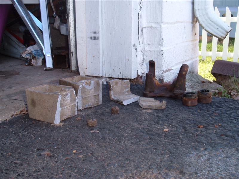 concrete blocks and rusty metal vehicle parts on asphalt near a white wall and downspout