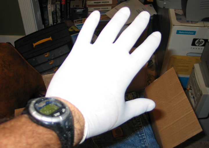 Hand wearing a white glove and a digital watch, with background clutter including boxes and a toolbox