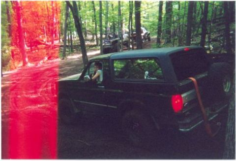 1996 Ford Bronco - A black 1996 Ford Bronco drives through a wooded area, with other off-road vehicles visible in the background.