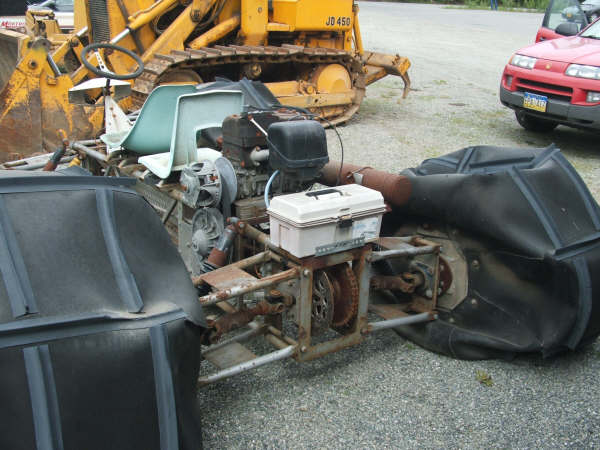 Partially assembled vehicle with engine, large black tires, and a toolbox on top. Yellow bulldozer and red car in background