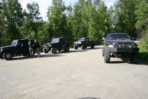 2003 Jeep TJ - Several modified off-road vehicles are parked on a dirt road, with a dense forest visible behind them.