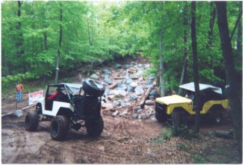 1996 Ford Bronco - Two open-top Jeep CJ/Wrangler-style 4x4s staged at the bottom of a rocky trail obstacle in the woods, a white one with a red roll bar, rear…