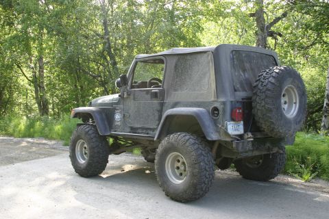 2003 Jeep TJ - A dusty Jeep Wrangler is parked on a dirt road surrounded by trees, with a spare tire mounted on the back.