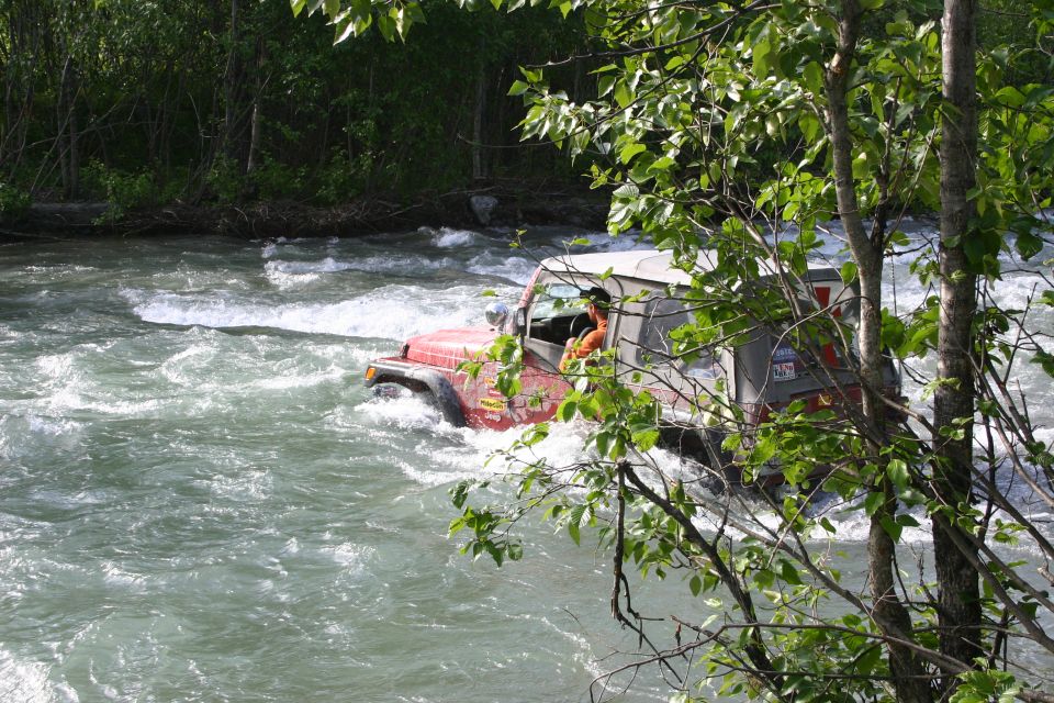 Red Jeep Wrangler driving through a river, driver visible, trees in foreground