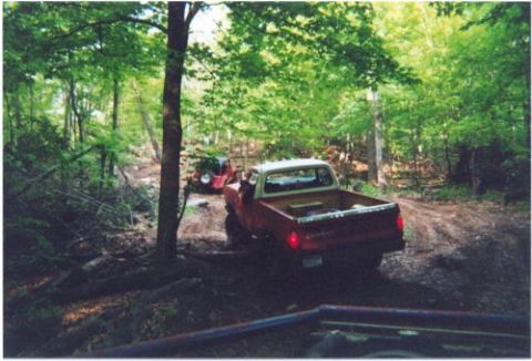 1996 Ford Bronco - Rear three-quarter view of a red vintage pickup with a white roof and step-side style bed working down a rutted dirt trail through green wo…