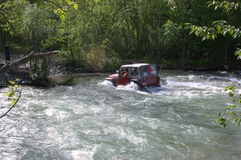 2003 Jeep TJ - A red Jeep Wrangler navigates a rocky river, with lush green trees lining the banks.