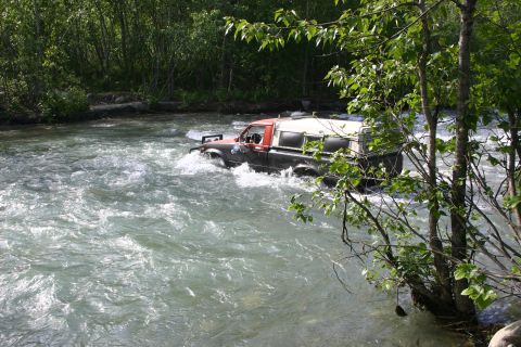 2003 Jeep TJ - A modified pickup truck navigates a rocky riverbed surrounded by lush green foliage.