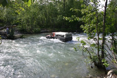 2003 Jeep TJ - An off-road vehicle navigates a fast-flowing river surrounded by lush green trees.
