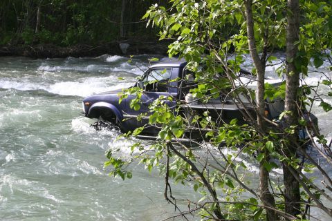 2003 Jeep TJ - A blue pickup truck navigates a rough river, its tires submerged in the turbulent water.