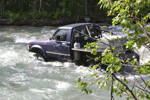 2003 Jeep TJ - A modified blue pickup truck drives through a fast-moving river, its off-road capabilities evident.