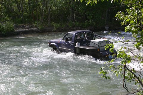 2003 Jeep TJ - Pickup truck navigates a river, with trees and foliage visible on the banks.