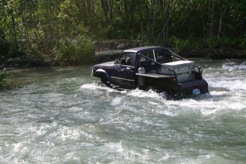 2003 Jeep TJ - A dark blue pickup truck navigates a shallow river with trees lining the banks.