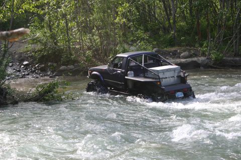 2003 Jeep TJ - A dark pickup truck navigates a rocky riverbed, its large tires churning through the water.