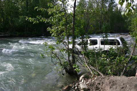 2003 Jeep TJ - White pickup truck stuck in a river, with trees and foliage on the banks.