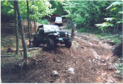 1996 Ford Bronco - A green square-headlight Jeep Wrangler YJ with a white soft top folded back, front bumper lights and aggressive mud tires crawls down a rut…