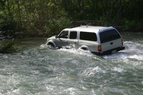 2003 Jeep TJ - A silver pickup truck navigates a rocky riverbed, its tires submerged in the rushing water.