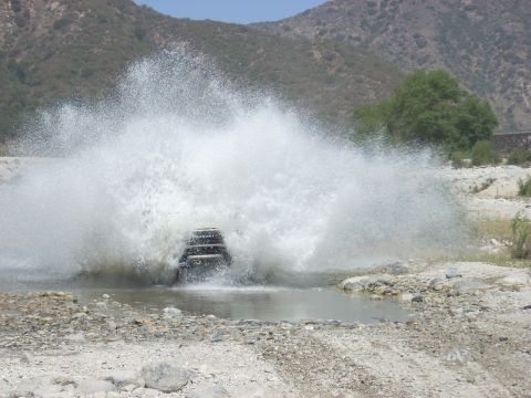1987 Suzuki Samurai - A dark SUV creates a large splash as it drives through a shallow riverbed.