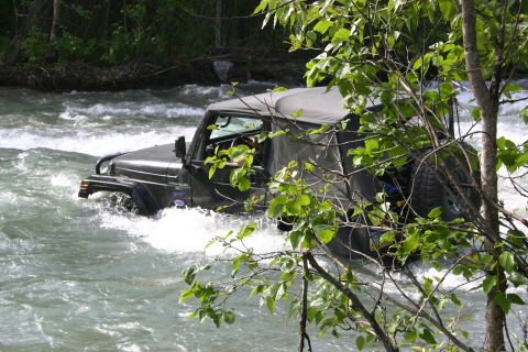 2003 Jeep TJ - A Jeep Wrangler navigates through a fast-moving river, surrounded by lush green foliage.