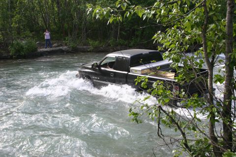 2003 Jeep TJ - A black pickup truck navigates a river, with a person standing on the bank in the background.