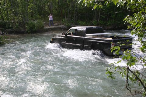 2003 Jeep TJ - A black pickup truck navigates through a fast-moving river, with a person watching from the bank.